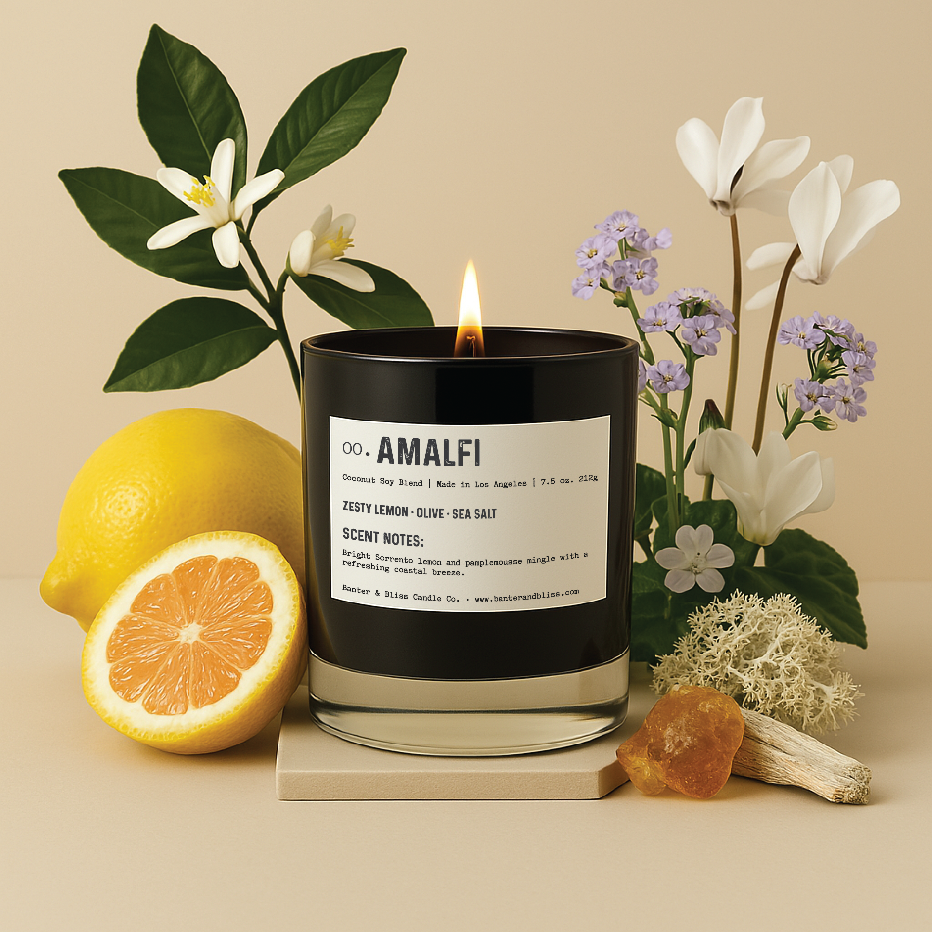 A lit AMALFI candle stands on a beige platform next to a halved lemon, citrus blossoms, cyclamen flowers, driftwood, and amber resin. The background features leafy greenery and blooming flowers in soft lighting.