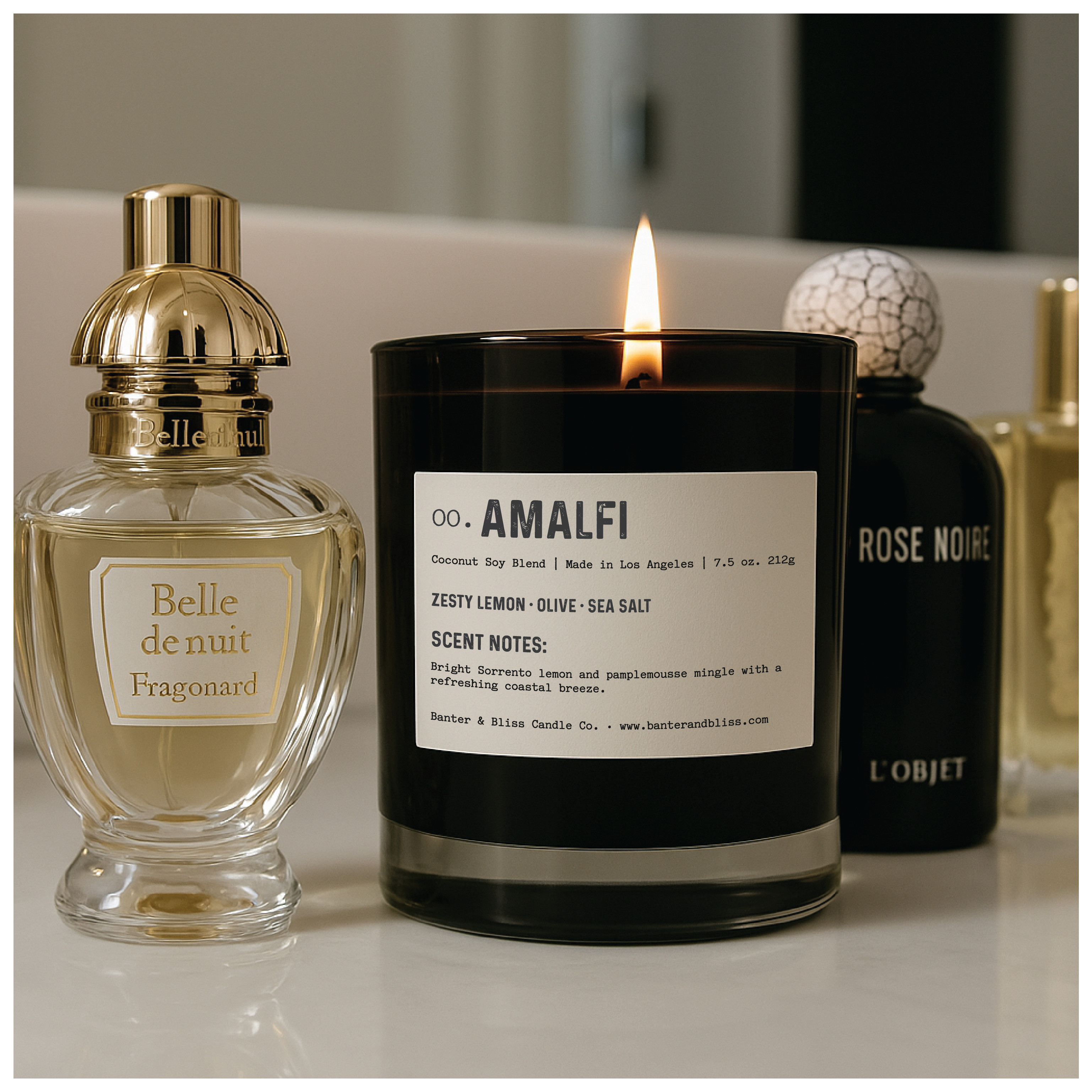 A black AMALFI candle burns on a bathroom vanity surrounded by luxury perfume bottles, including Belle de Nuit by Fragonard and Rose Noire by L’Objet. The candle’s white label displays scent notes and branding details.