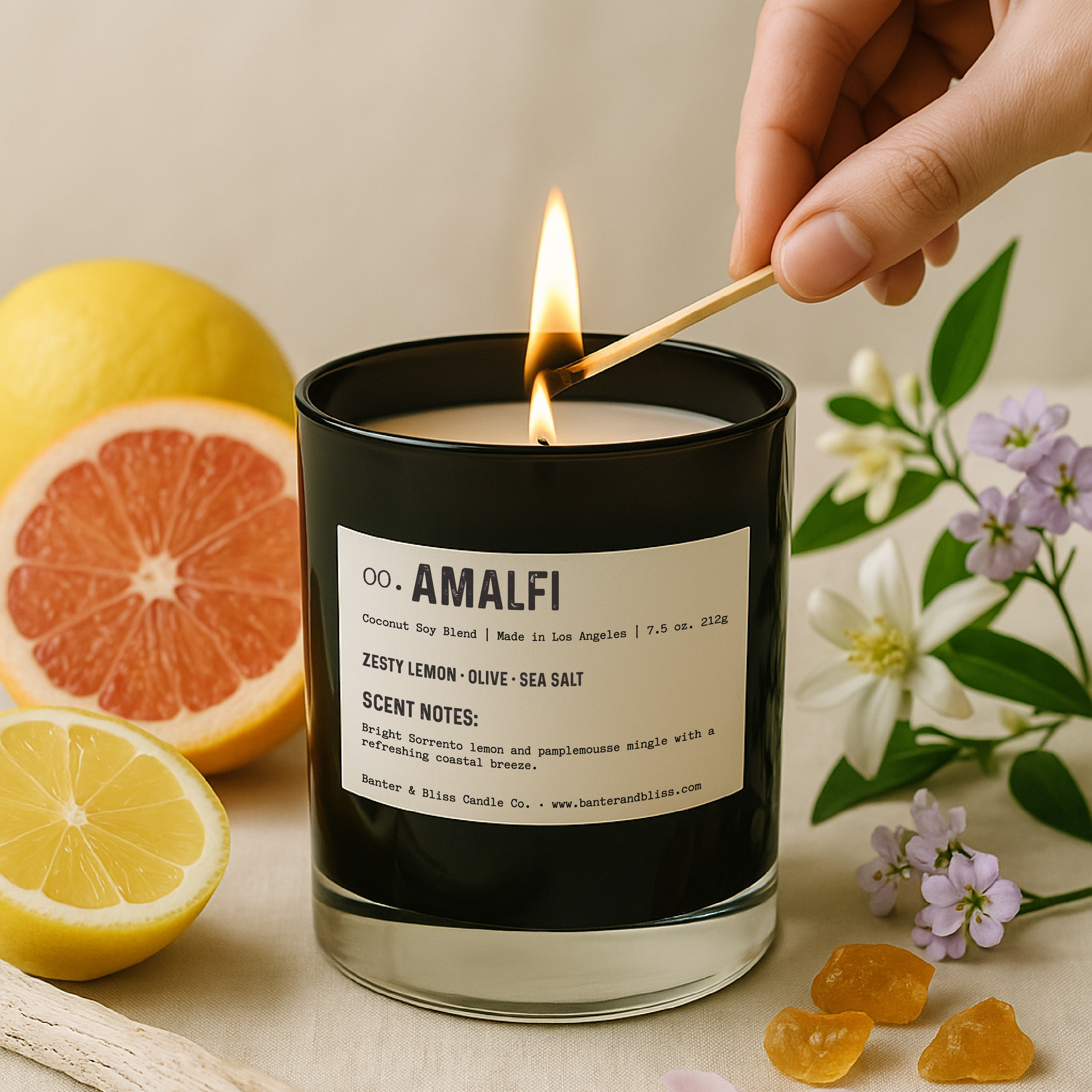 A hand is seen lighting a black glass candle labeled "00. AMALFI" by Banter & Bliss Candle Co. The candle sits on a light fabric surface surrounded by sliced grapefruit and lemon, white and purple flowers, amber resin, and a piece of driftwood. The candle label lists scent notes including zesty lemon, olive, and sea salt. The overall aesthetic is bright, natural, and styled in a soft, professional tone.