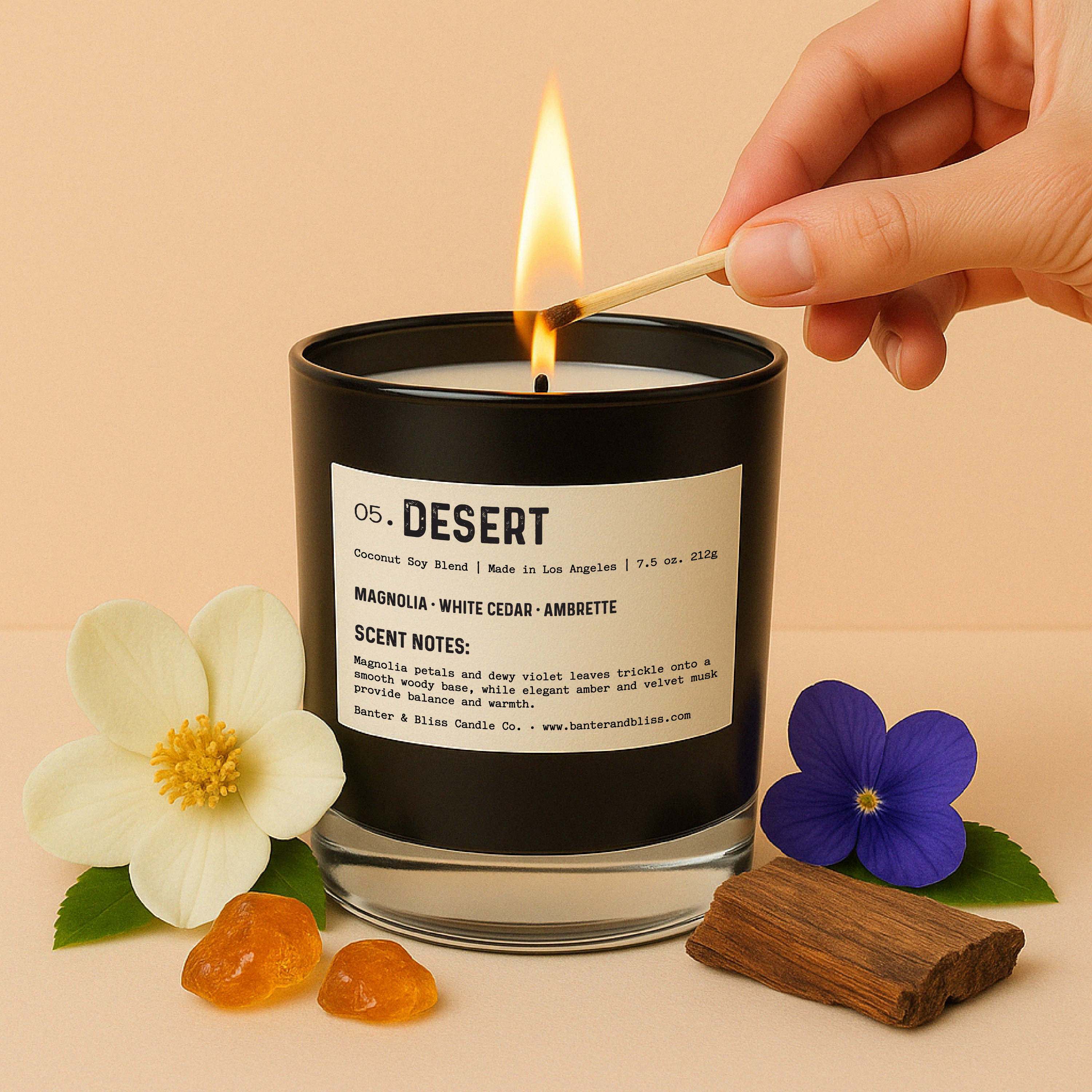 Lit black candle labeled “05. DESERT” surrounded by realistic magnolia flowers, violet blossom, green leaves, amber resin pieces, and a piece of cedar wood. Set against a soft pastel background for a warm, natural aesthetic.