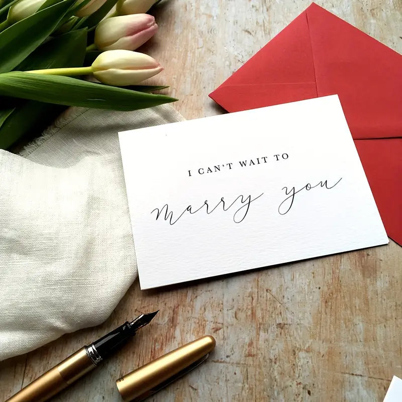 A flat lay image of a romantic card on a wooden surface. The card has elegant, minimal black script that reads "I CAN'T WAIT TO marry you." It is paired with a red envelope, a gold fountain pen, a soft cream-colored cloth, and a bouquet of white and pale pink tulips, creating a warm and heartfelt atmosphere.
