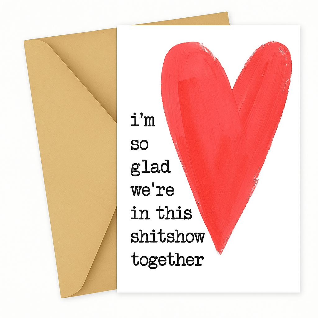 A vertical white greeting card placed on top of a kraft brown envelope, shown against a clean white background. The card features a large, hand-painted red heart on the right and black typewriter-style text on the left that reads: "i'm so glad we're in this shitshow together."
