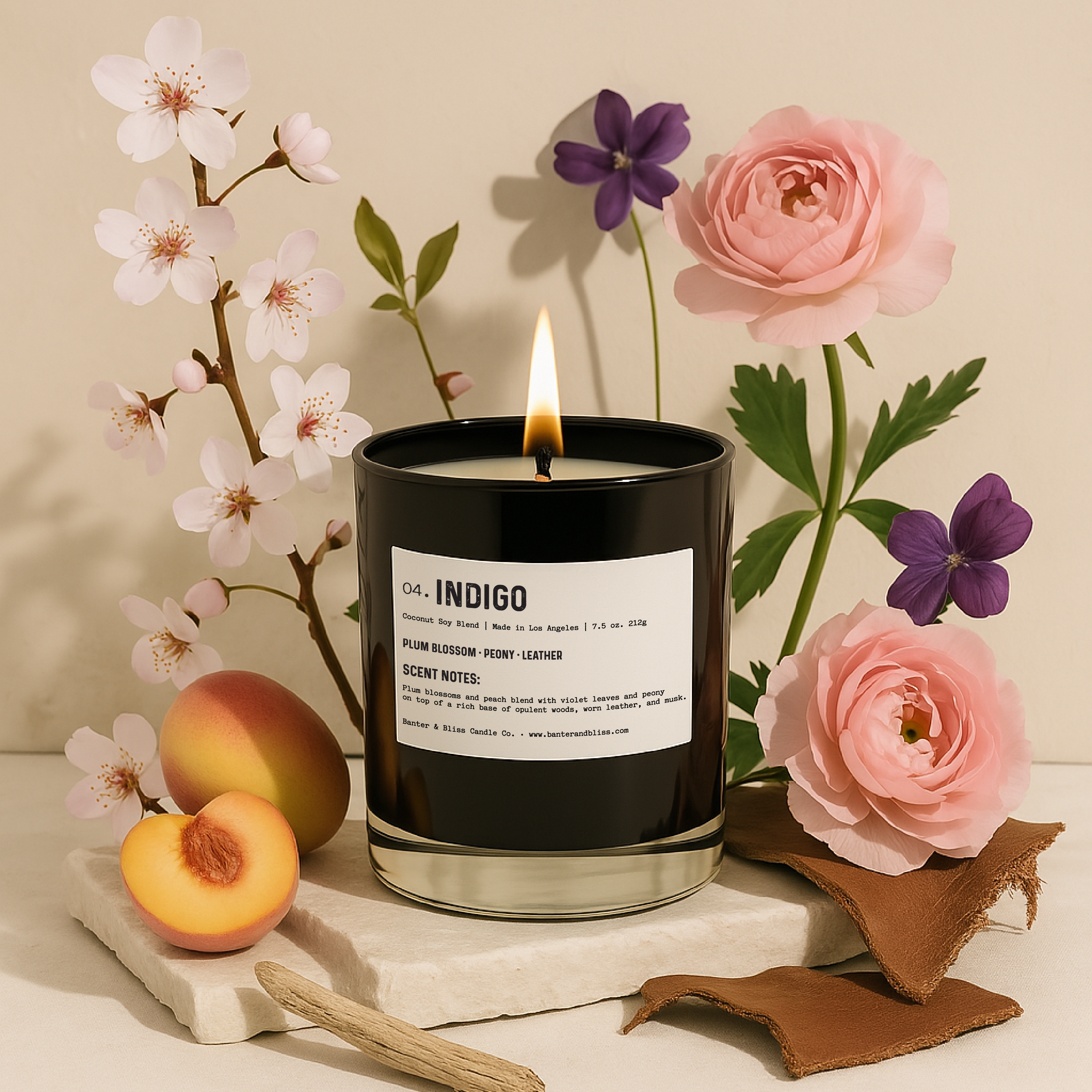 A lit black glass candle labeled "04. INDIGO" by Banter & Bliss Candle Co. is surrounded by elements that evoke its fragrance notes: pink ranunculus, plum blossoms, purple violet flowers, soft peach slices, driftwood, and pieces of worn leather. The candle sits on stacked stone tiles against a neutral cream backdrop.