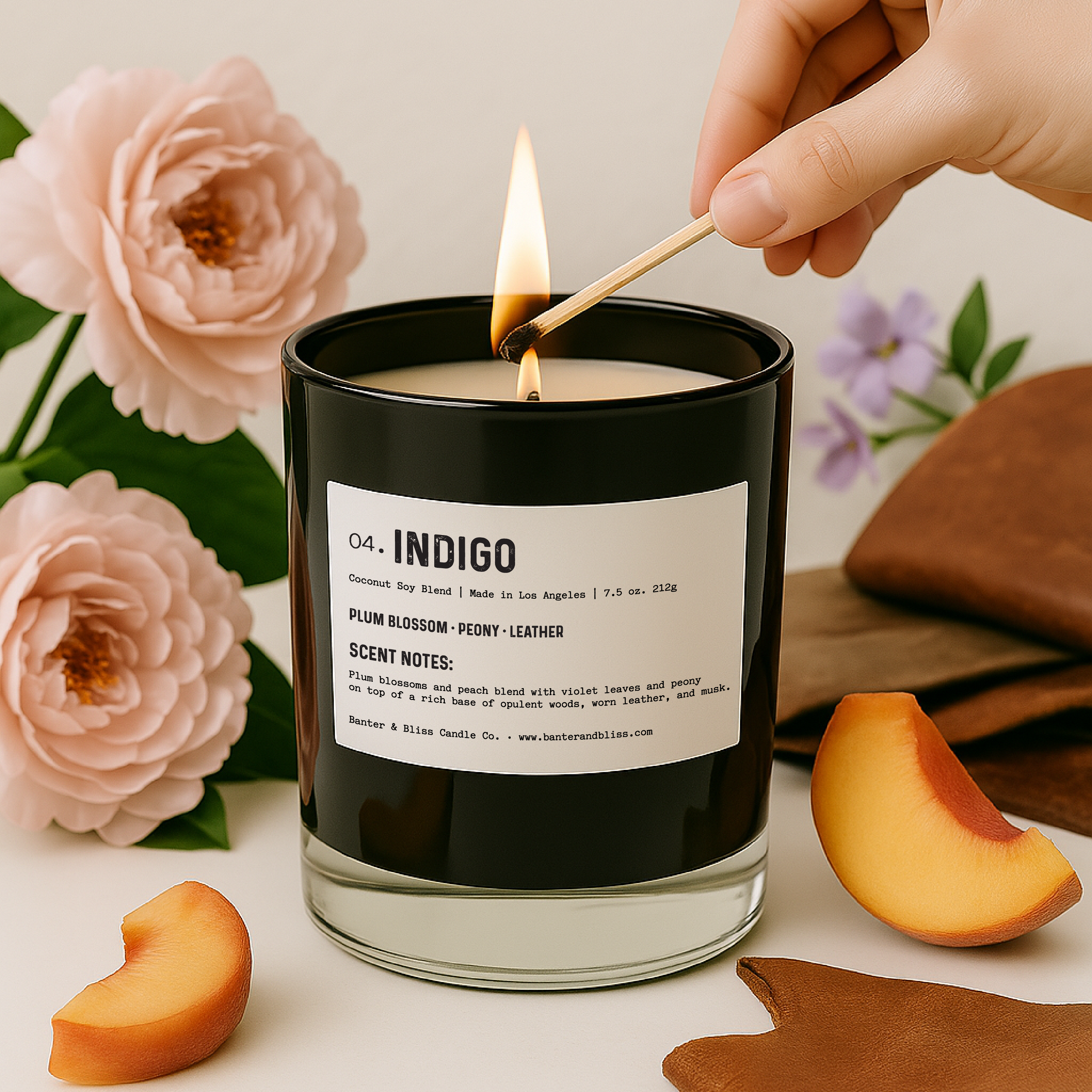A hand is seen lighting a black glass candle labeled "04. INDIGO" by Banter & Bliss Candle Co. The scene includes soft peach slices, a folded piece of brown leather, and blooming peonies and violet flowers in the background, styled against a clean, white surface in a natural light setting.