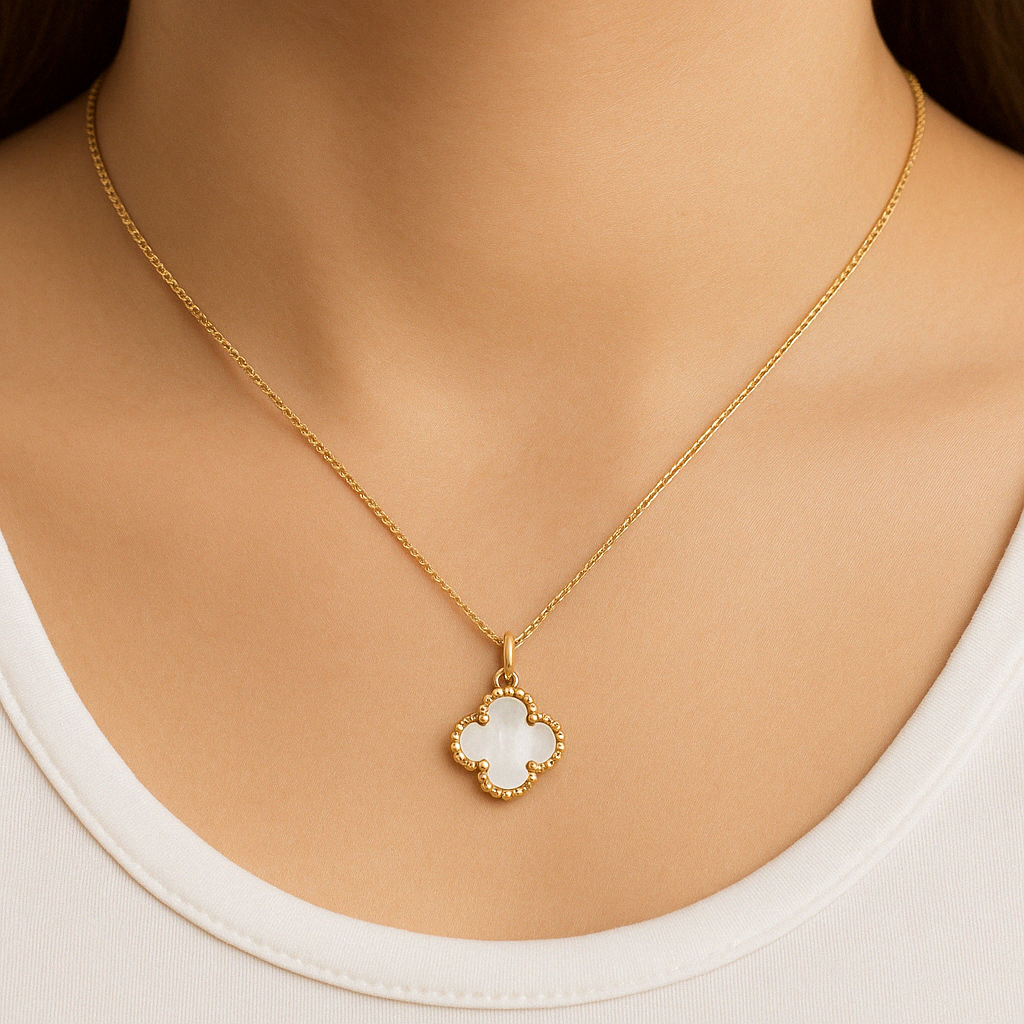 A close-up photo of a woman wearing a gold necklace with a clover-shaped mother-of-pearl pendant. The necklace rests elegantly against her skin just below the collarbone, with the pendant centered and the chain forming a soft curve around her neck.