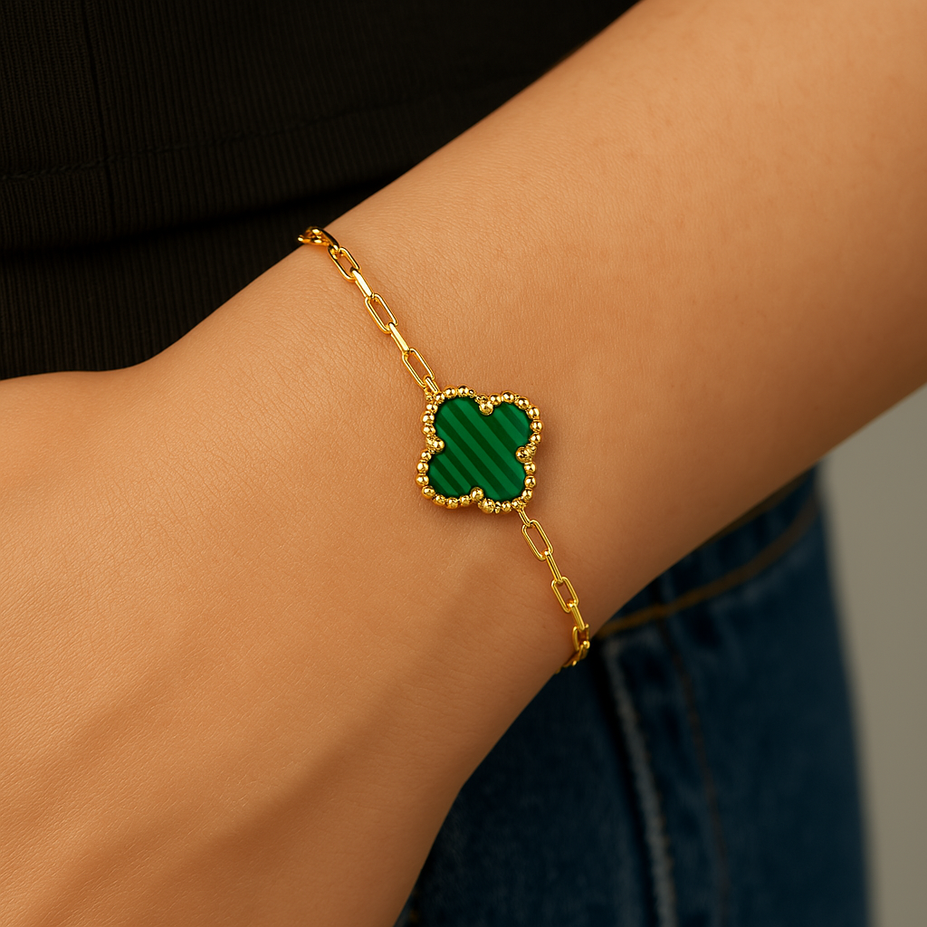 A lifestyle photo showing a gold bracelet with a clover-shaped malachite charm worn on a person’s wrist. The background includes part of a black top and blue denim jeans, and the green-striped malachite and yellow gold chain are clearly visible in natural lighting.