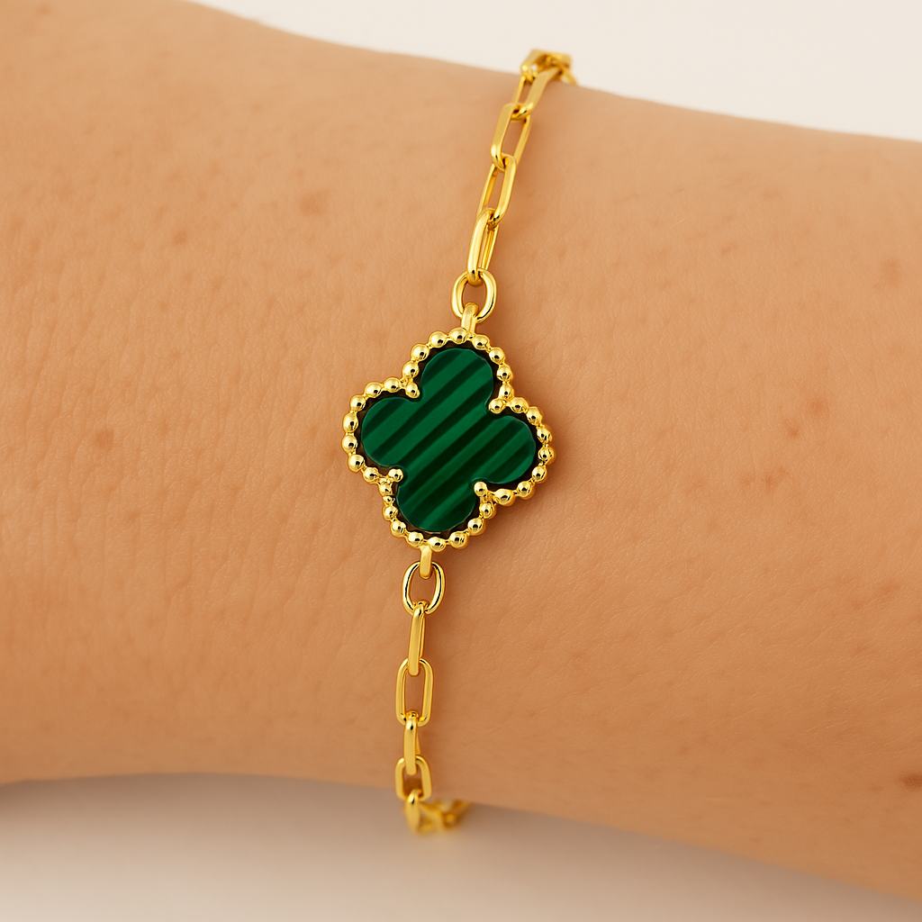 A close-up image of a gold bracelet with a clover-shaped malachite pendant resting on a person’s wrist. The malachite stone has dark green stripes and is framed with a beaded gold border, highlighted under soft lighting.