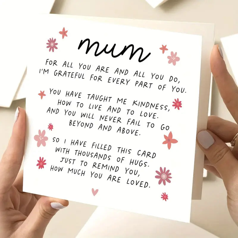 Close-up of a person holding a square white greeting card with pink flower illustrations and a heartfelt message in black cursive and print text. The card begins with the word 'mum' and expresses gratitude, love, and appreciation for a mother's kindness and support. The person's hands have light-colored nail polish and are gently holding the card in a well-lit setting.