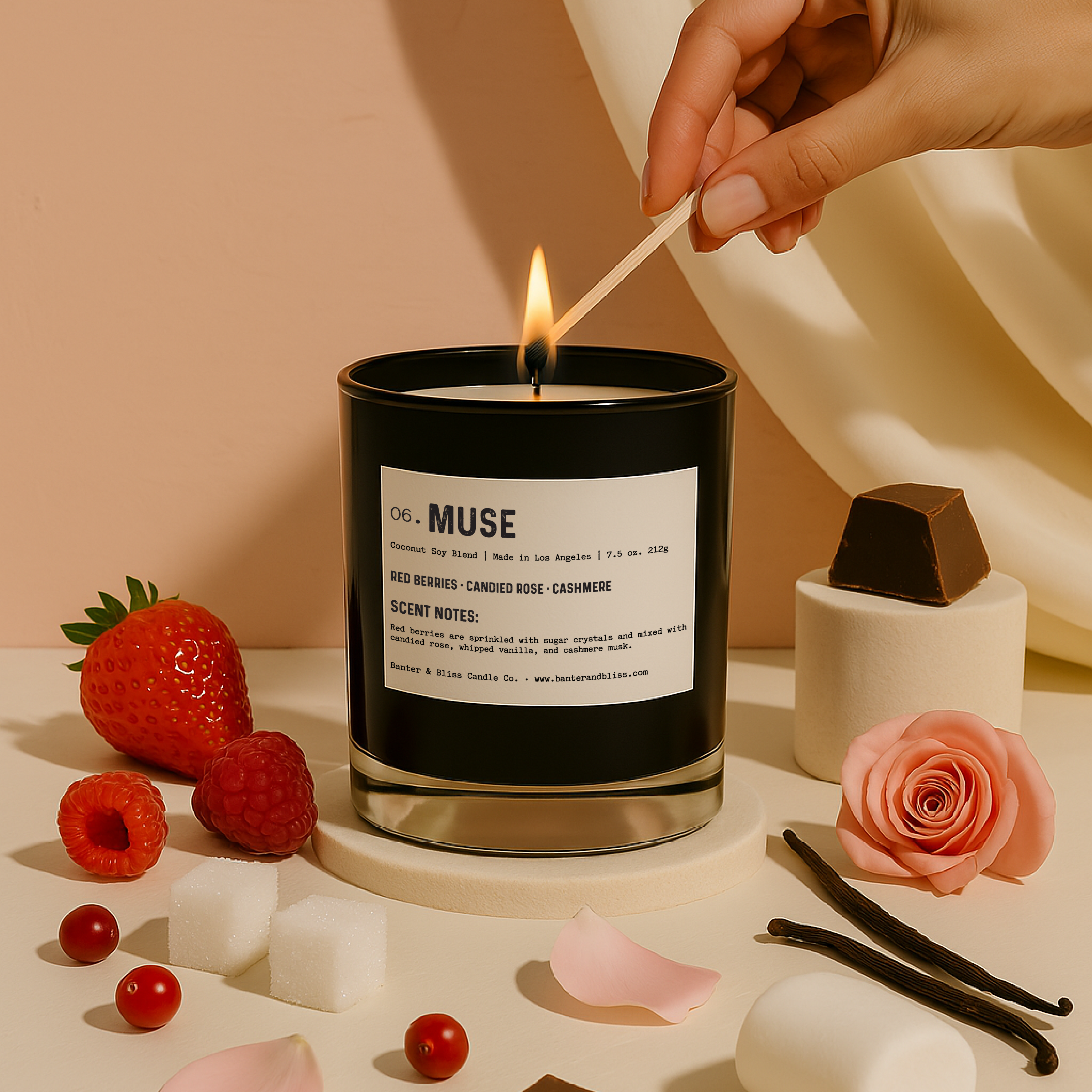 A luxury candle labeled "06. MUSE" is being lit by a hand holding a matchstick. The black glass candle is centered on a circular beige pedestal, surrounded by representative scent notes including fresh strawberries, raspberries, sugar cubes, red berries, chocolate, rose petals, a full rose, vanilla beans, and a marshmallow. The background features a peach-toned wall and soft cream drapery, evoking an elegant and warm aesthetic.