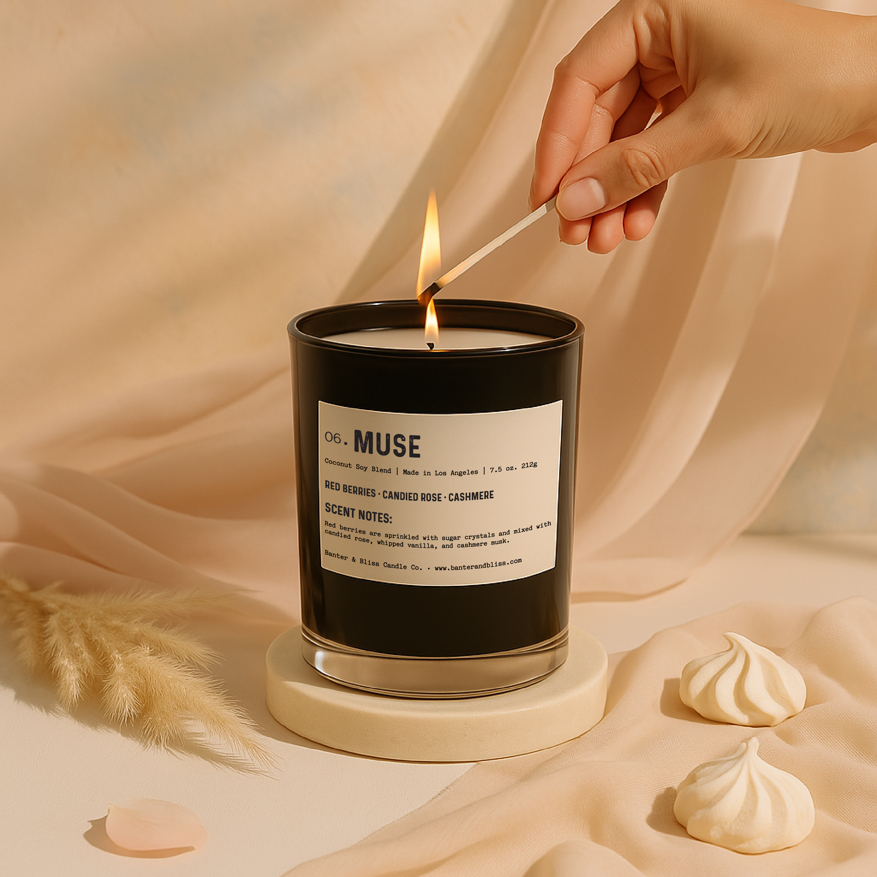 A luxury candle labeled "06. MUSE" is being lit by a hand holding a match. The black candle is placed on a cream-colored pedestal with soft beige fabric draped in the background. Surrounding the candle are delicate decorative elements including a pink rose petal, fluffy pampas grass, and vanilla meringue cookies. The scene is warmly lit, creating an elegant and calming ambiance suited to the candle’s scent notes of red berries, candied rose, whipped vanilla, and cashmere musk.