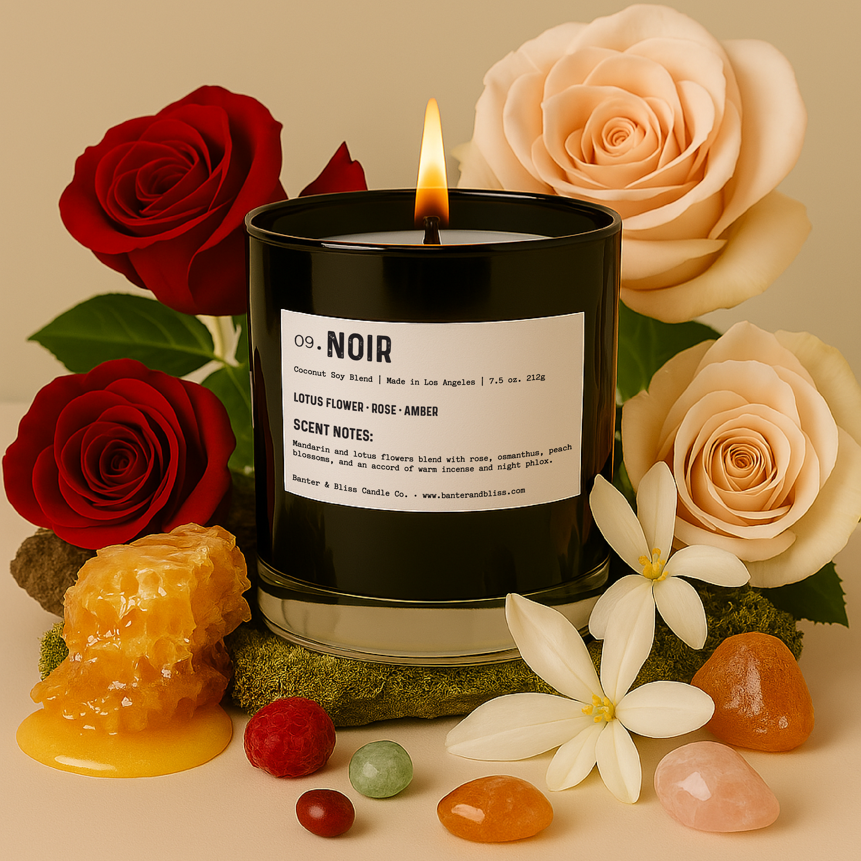 A lit black candle labeled “09. Noir” sits surrounded by red and blush roses, white jasmine flowers, amber resin, honeycomb dripping with honey, and colorful polished stones. The scene is styled on a soft beige surface with green moss, creating a luxurious and earthy aesthetic.