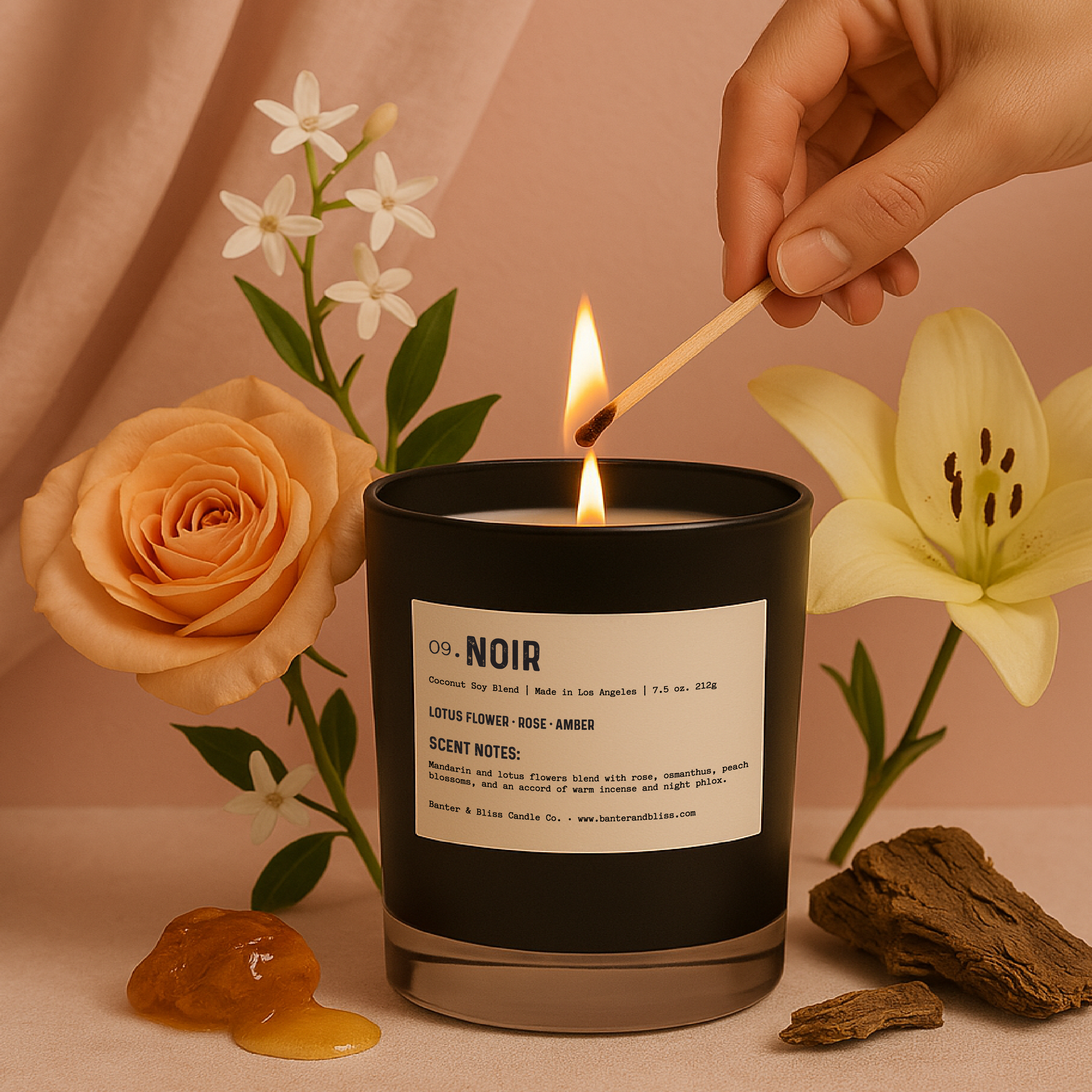 A luxury black candle labeled “09. Noir” is being lit by a hand holding a wooden match. The candle is surrounded by a peach rose, white jasmine, a yellow lily, amber resin, and honey on a soft blush pink surface. A blush curtain and wall form the background, evoking a warm and elegant atmosphere.