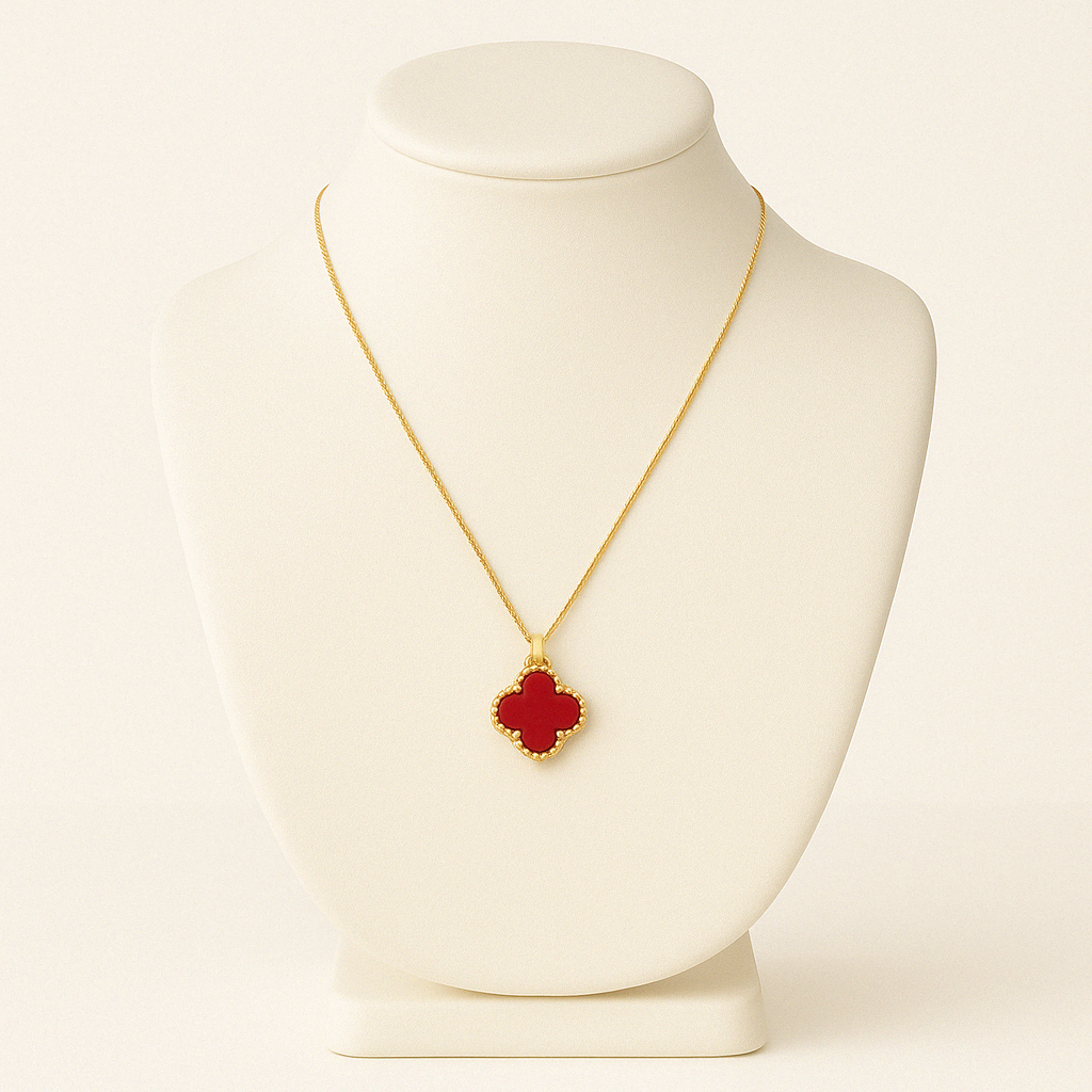 Gold necklace with a red clover-shaped pendant displayed on a smooth white jewelry bust, set against a white background. The pendant features a glossy red enamel center bordered by a delicate gold beaded frame, with soft lighting highlighting the chain and pendant details.