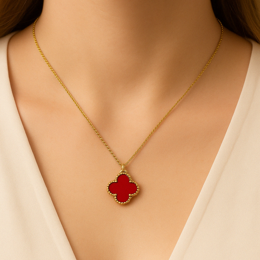 Close-up view of a woman's neck wearing a gold necklace with a red clover-shaped pendant, centered below her collarbone. The pendant has a smooth red surface framed by a beaded gold border, and the model is dressed in a cream-colored blouse.