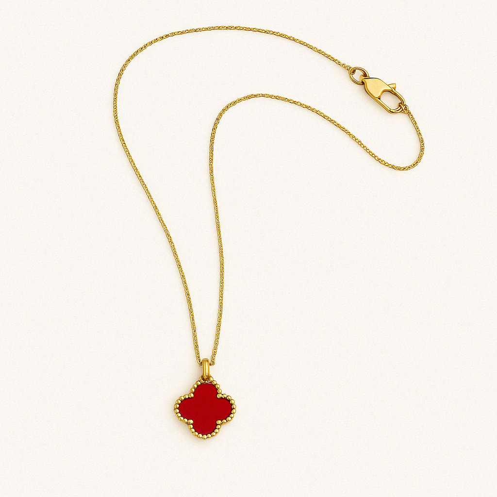 Gold chain necklace with a red clover-shaped pendant outlined in a beaded gold frame, elegantly laid out on a white background with a soft curve in the chain and the clasp visible in the top corner.