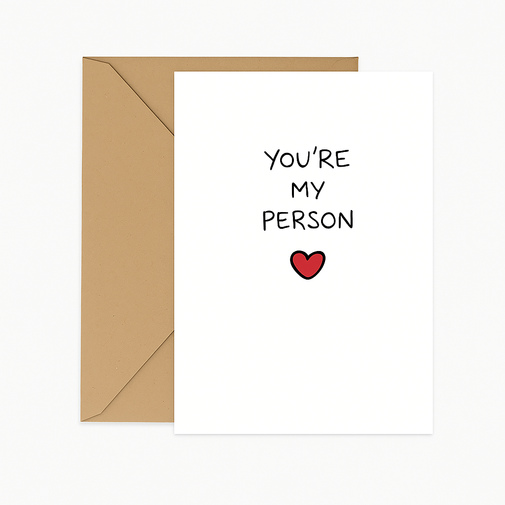 A white greeting card placed on top of a brown kraft envelope, shown against a clean white background. The card features black handwritten-style text that reads, "YOU'RE MY PERSON," with a small red heart illustration centered below the text.
