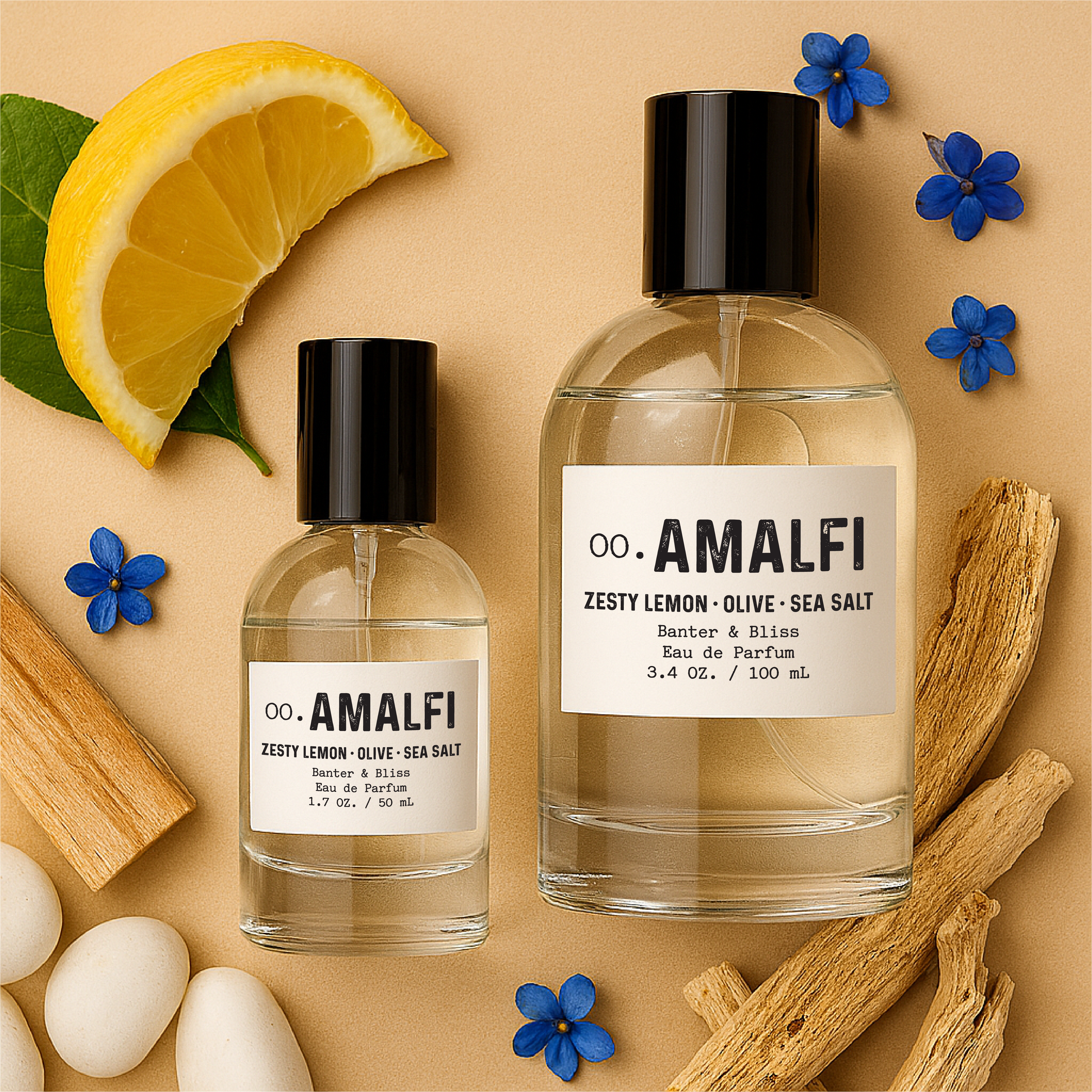Flatlay of Banter & Bliss "Amalfi" Eau de Parfum bottles in 1.7 oz and 3.4 oz sizes, surrounded by lemon slices, blue cyclamen flowers, white driftwood, and smooth stones on a pastel beige background — fragrance notes include zesty lemon, olive, sea salt, citrus blossom, and amber mist.