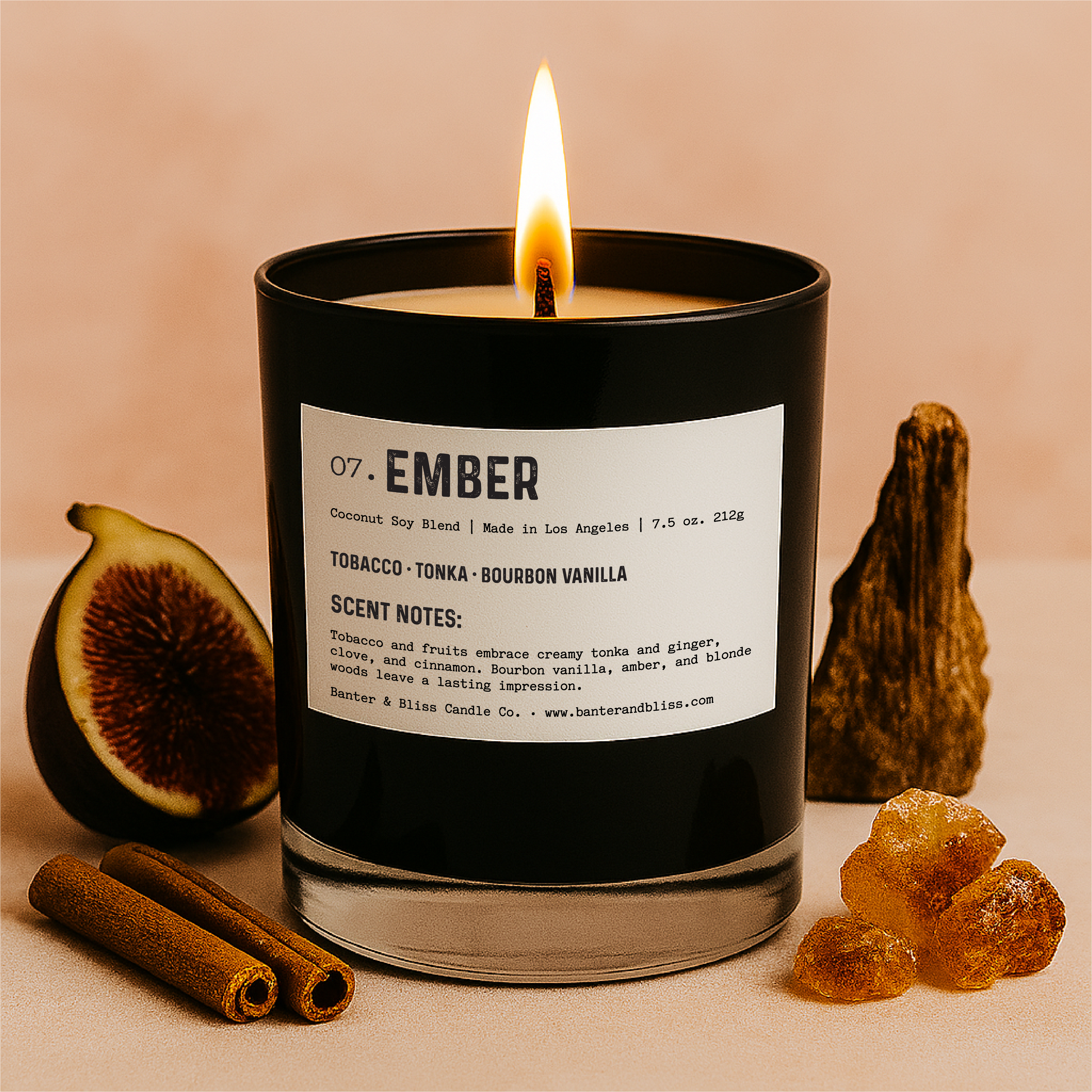 Ember candle by Banter & Bliss Candle Co. in black glass jar with label, lit and styled with figs, cinnamon sticks, amber crystals, and driftwood on a pastel peach background — features scent notes of tobacco, tonka, and bourbon vanilla.