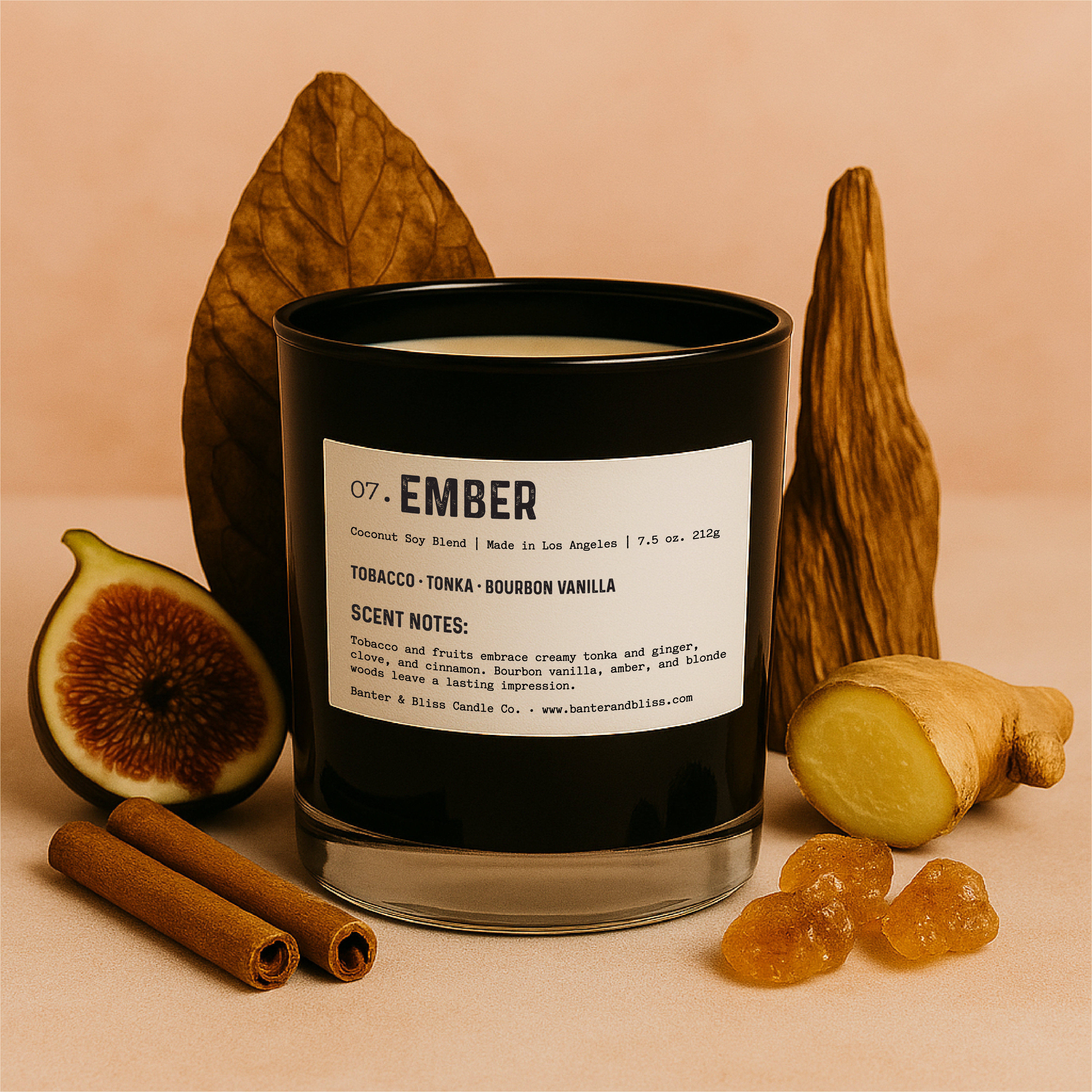 Banter & Bliss "Ember" candle in a matte black glass jar with product label, surrounded by sliced fig, cinnamon sticks, ginger root, amber crystals, and dried tobacco leaf, set against a pastel peach background – featuring scent notes of tobacco, tonka, bourbon vanilla, and warm spices.