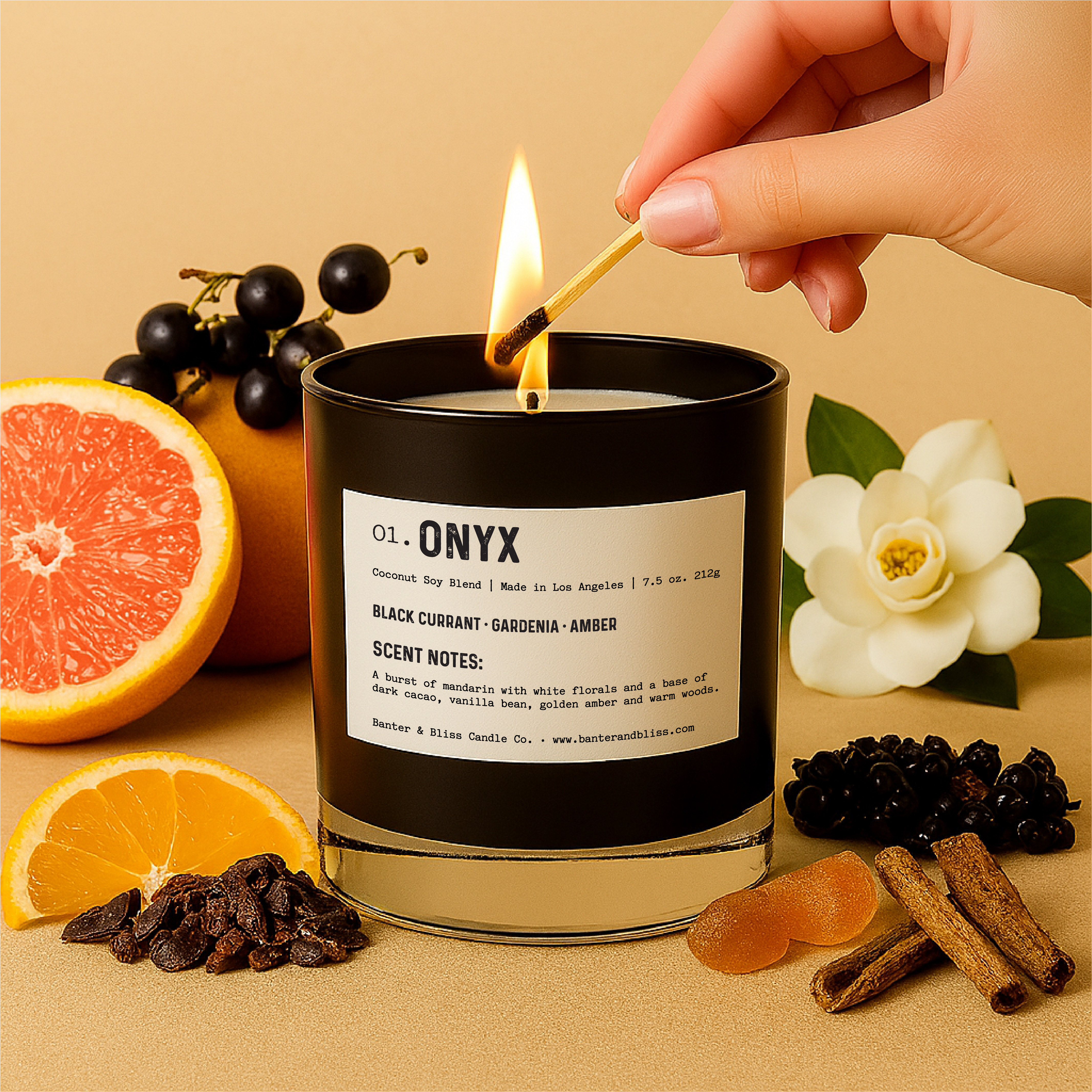 Banter & Bliss "Onyx" candle in a black glass jar being lit with a match, surrounded by black currants, mandarin orange slices, cacao nibs, white gardenia, vanilla bean, golden amber, and sandalwood — set against a pastel beige background. Features scent notes of black currant, gardenia, amber, dark cacao, and creamy woods.