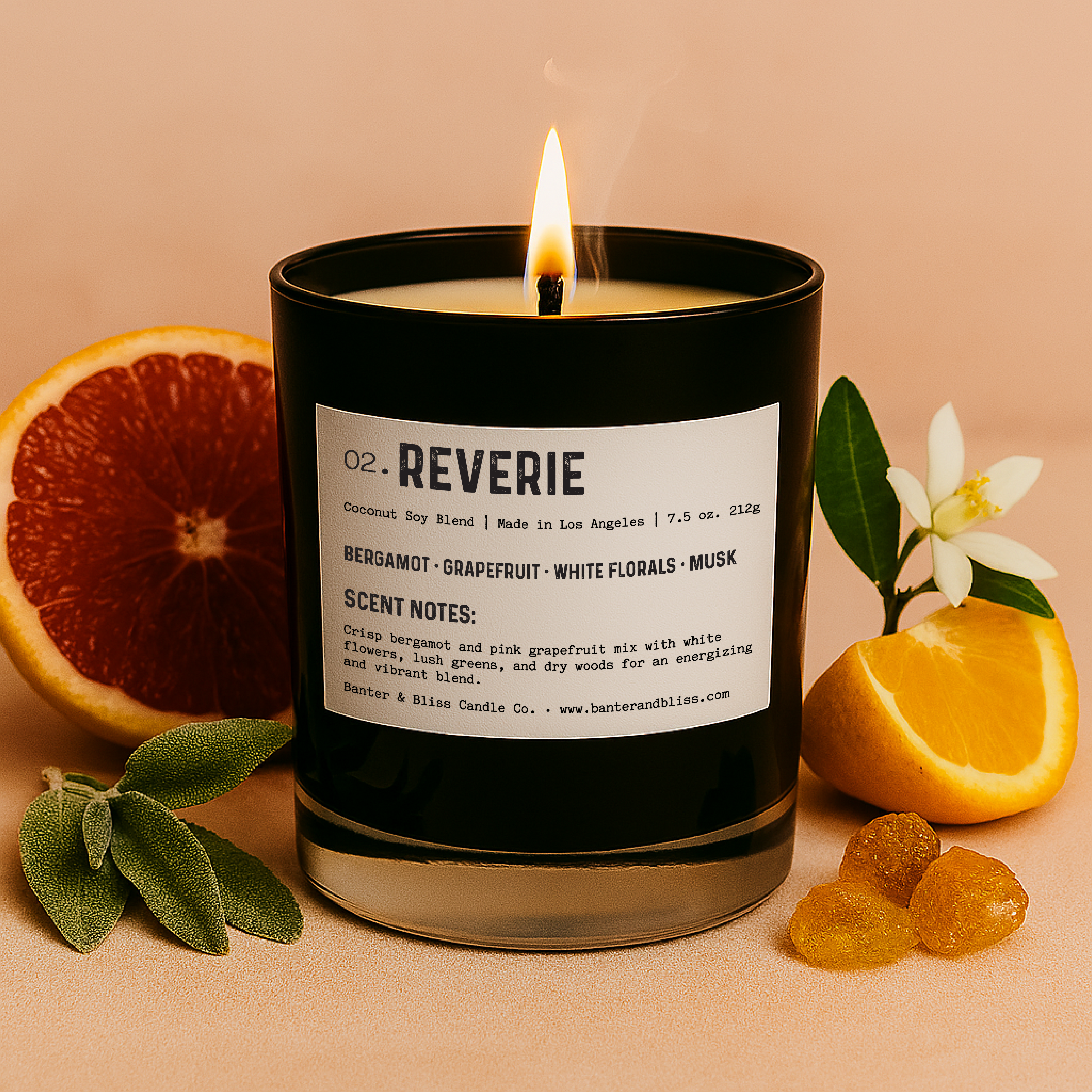 Banter & Bliss "Reverie" luxury candle in black glass jar with product label, lit and styled with fresh grapefruit, orange blossom, sage leaves, amber resin, and citrus on a pastel peach background — featuring scent notes of bergamot, grapefruit, white florals, musk, and dry woods.