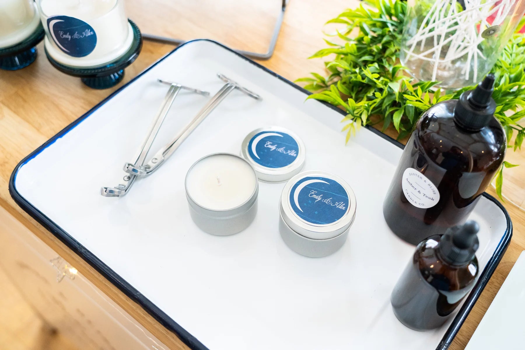 Candle making supplies on a white enamel tray, featuring Banter & Bliss branded travel tin candles, amber fragrance oil bottles, wick trimmers, and tools — DIY candle bar setup on wood table with greenery.