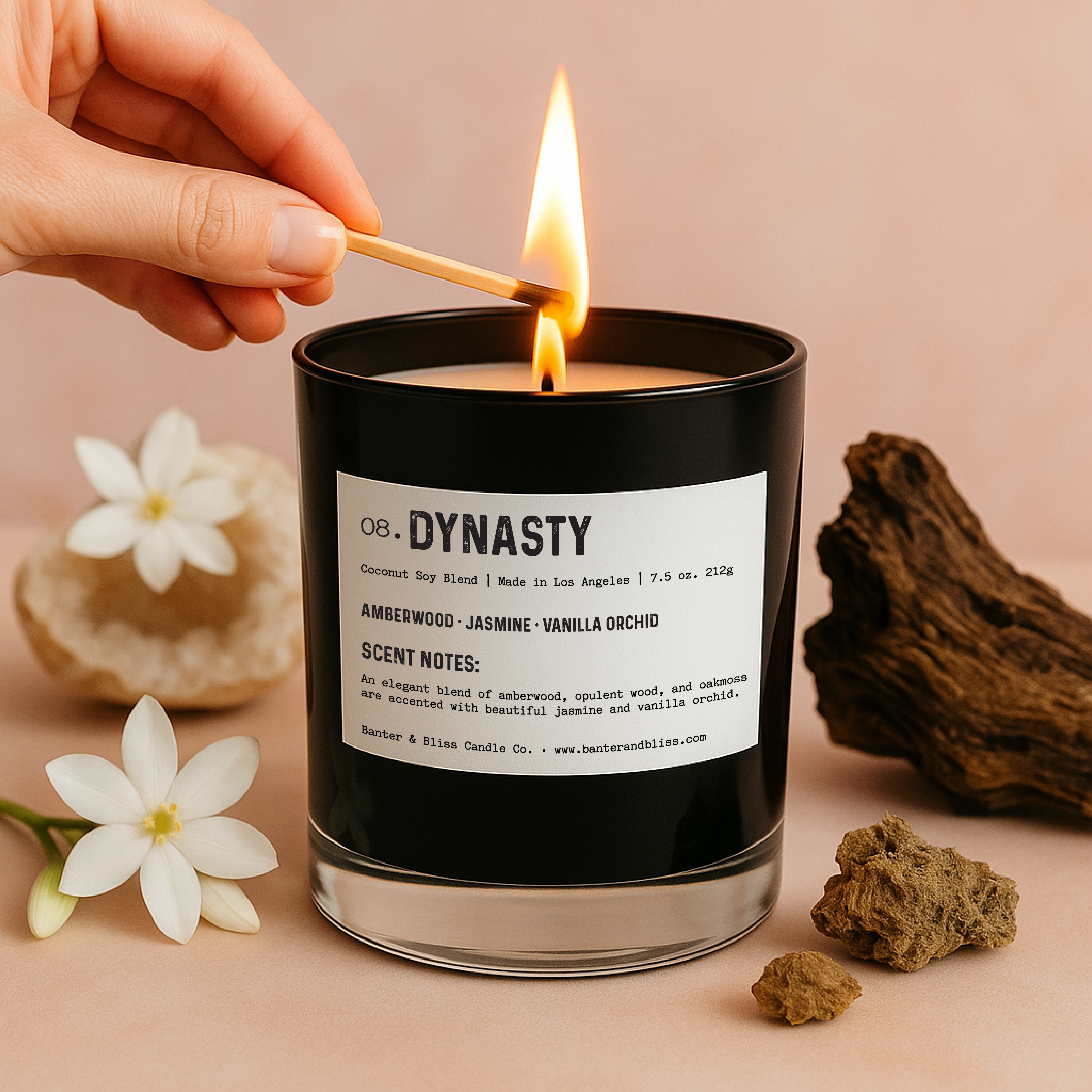 Luxury black candle labeled "Dynasty" by Banter & Bliss Candle Co. being lit with a match, surrounded by jasmine flowers, oakmoss, amberwood, and a pastel pink background – scented with amberwood, jasmine, and vanilla orchid.